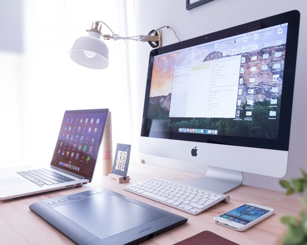 An iMac, MacBook Pro and Samsung mobile on with graphic tablet on a light wooden desk